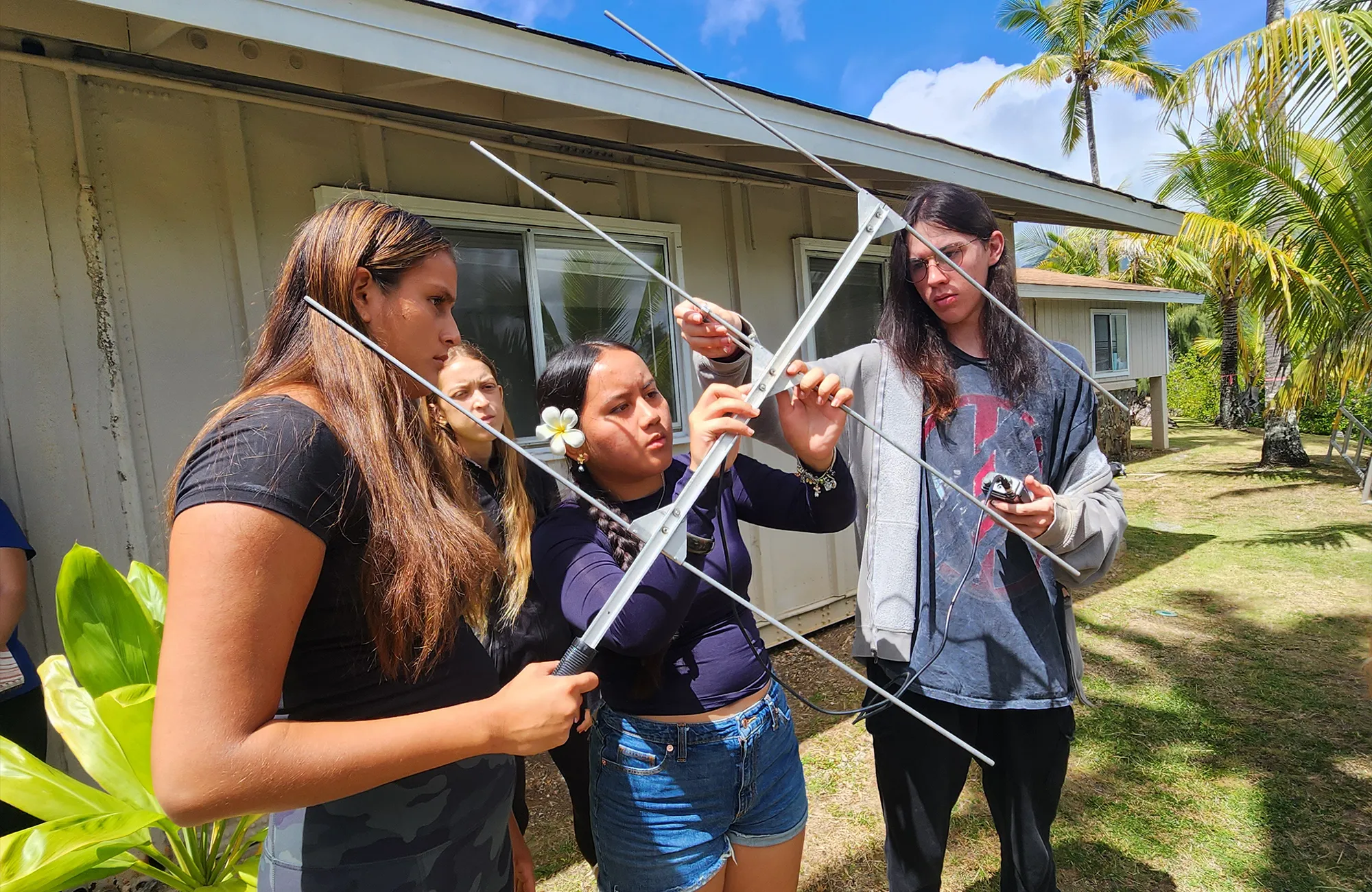 Three young people examine a handheld radio antenna outside near a building and palm trees on a sunny day.