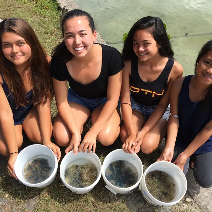 Four young women kneel on grass, each holding a white bucket filled with water and marine life near a shoreline.
