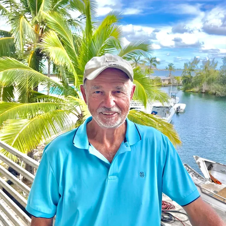 Smiling older man in a light blue polo and cap stands by palm trees and water with boats in the background.