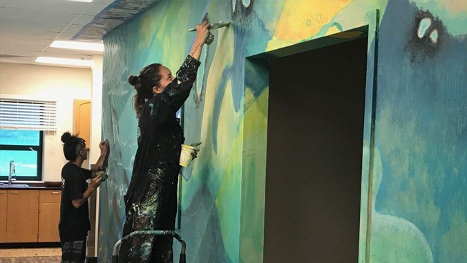 Two people paint a colorful mural on an indoor wall; one stands on a ladder while the other paints below.