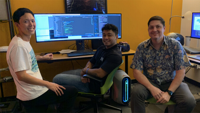 Three men smiling and sitting by a desk with a large computer monitor displaying code, in an office setting.