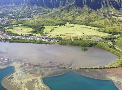 Aerial view of a coastal fishpond, green fields, and mountains in the background under clear skies.