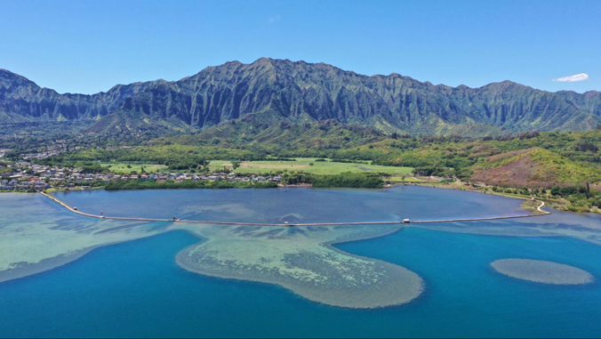Aerial view of a mountain range, lush greenery, a coastal lagoon, and a long bridge over blue water.