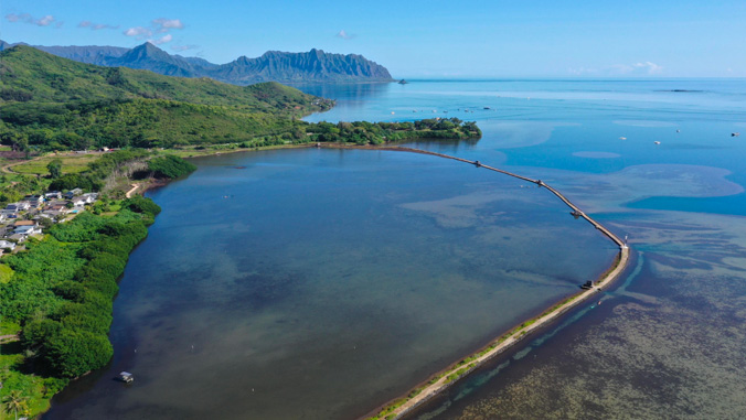 Aerial view of a long stone barrier crossing a calm bay, with mountains and a small village in the background.