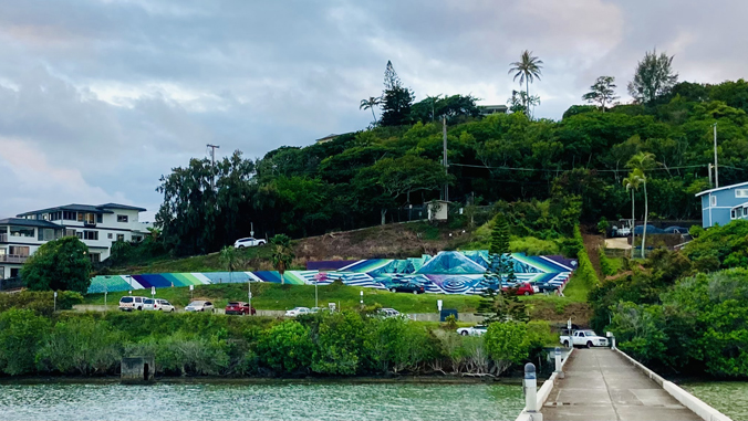 A colorful mural on a hillside overlooks a road, trees, houses, and a dock by the water under a cloudy sky.