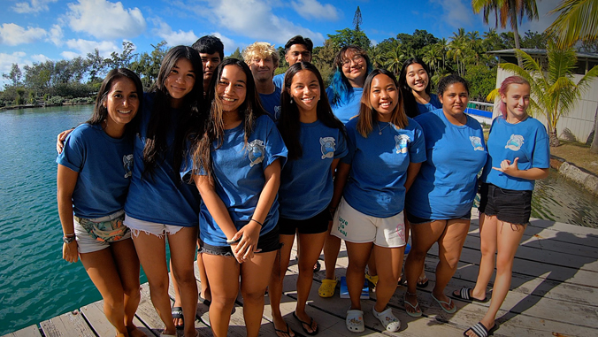 A group of smiling teens in blue shirts stand on a dock by the water with palm trees in the background.
