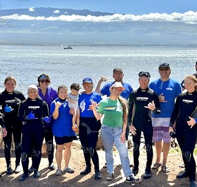 Group of people in wetsuits and casual clothes posing by the ocean with mountains in the background on a sunny day.