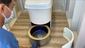Person wearing a mask removes a composting toilet waste container in a small bathroom with wood flooring.