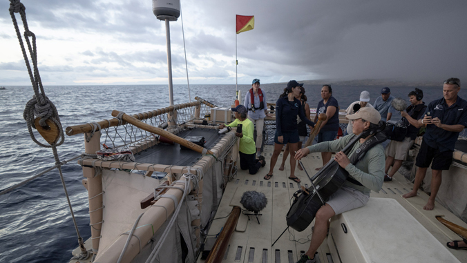 People aboard a canoe at sea; one person plays a cello while others observe under a cloudy sky.