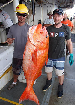 Two men on a boat proudly hold a large, bright orange fish that they have caught.
