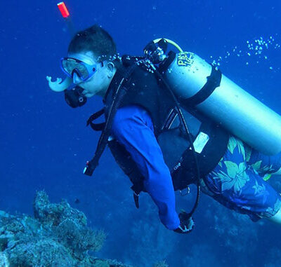 Person in scuba gear swimming underwater near coral, wearing a blue shirt and floral swim trunks.