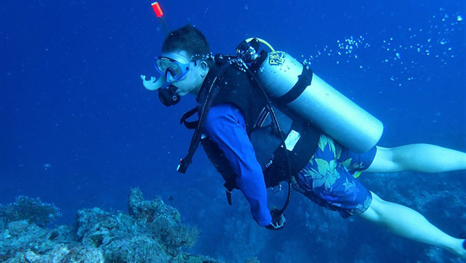 Person in scuba gear swimming underwater near coral, wearing a blue shirt and floral swim trunks.