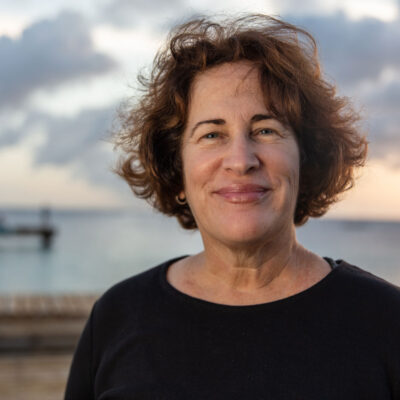 Woman with short brown hair smiling outdoors by the water at sunset, with a dock and cloudy sky in background.