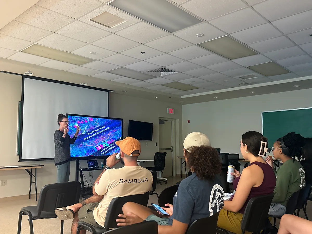 Person giving a presentation to a small group seated in a classroom, with a colorful slide on screen.