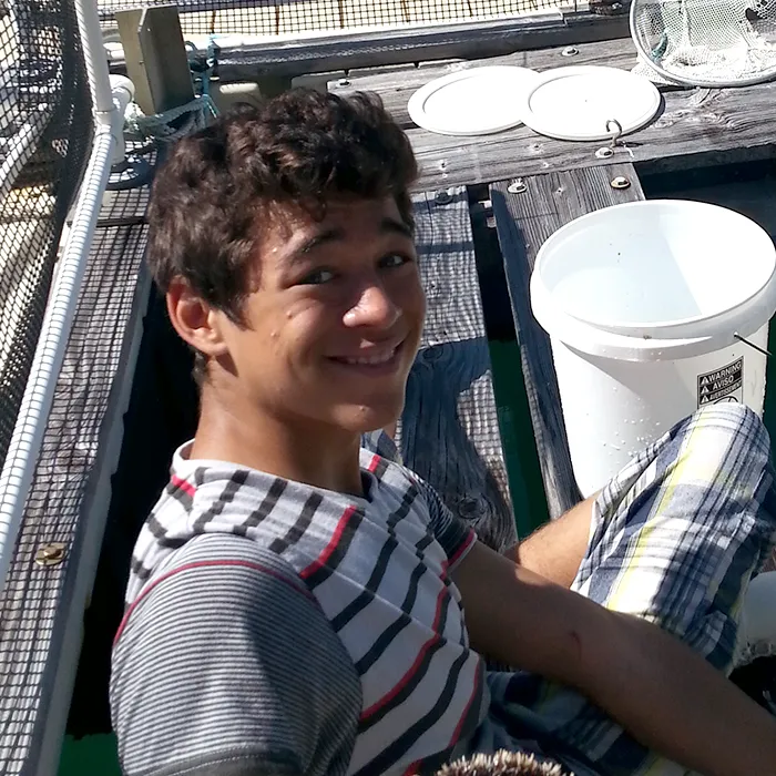 Teen boy smiling while sitting on a dock next to a white bucket on a sunny day.