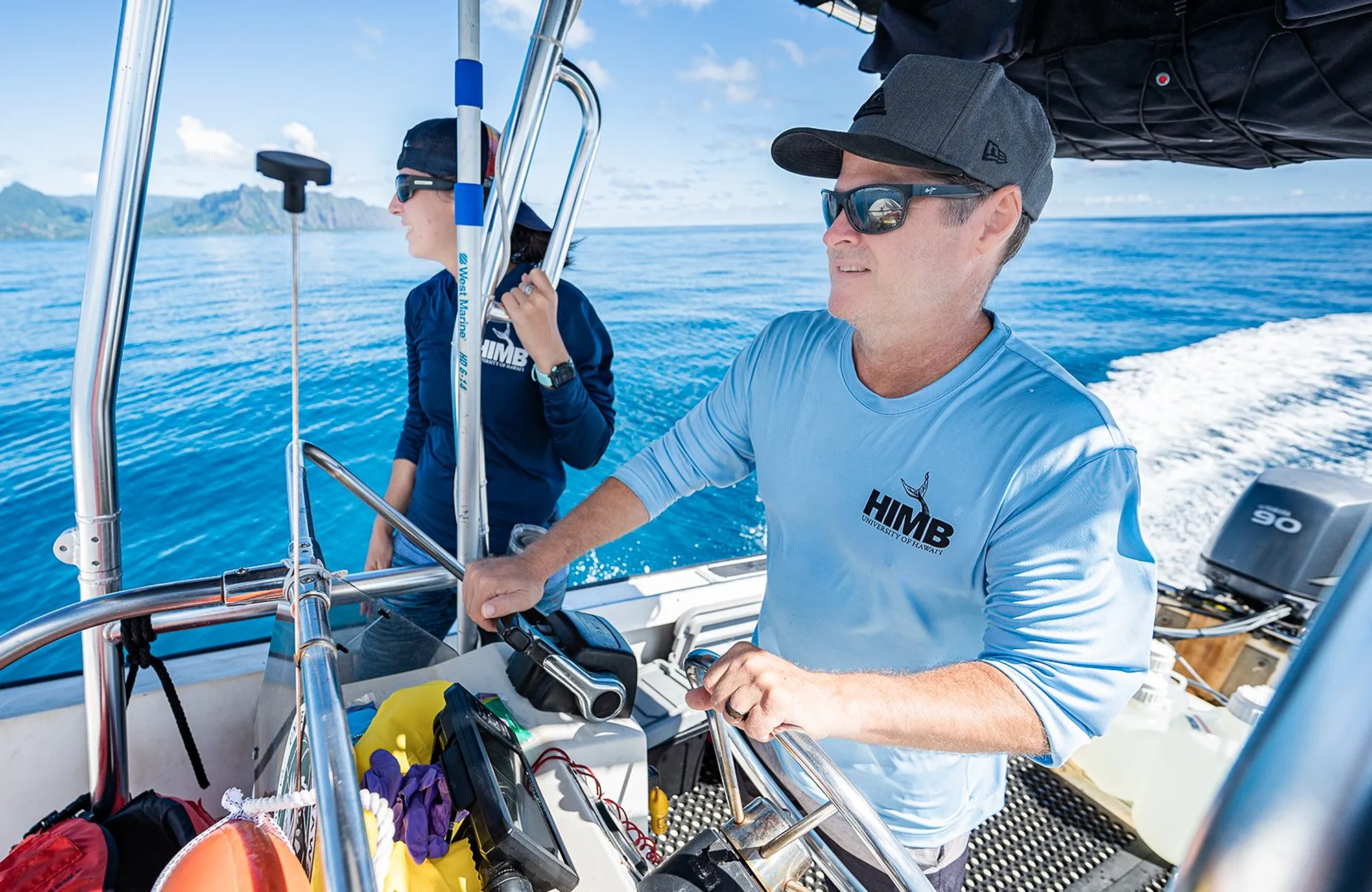 Two people steer a small research boat on clear blue water under a sunny sky, wearing hats and sunglasses.