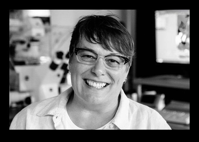 Smiling person with glasses and short hair in an office, shown in black and white.