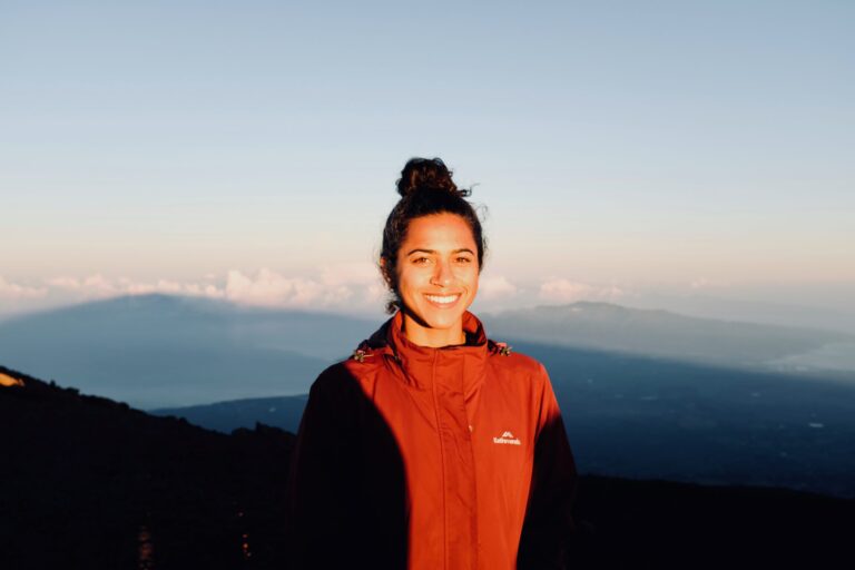 Woman in a red jacket smiles at sunrise with mountains and clouds in the background.