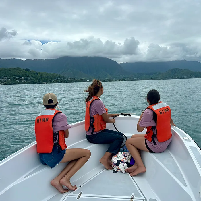 Three people in orange life vests sit on a boat, looking at mountains under a cloudy sky on the water.