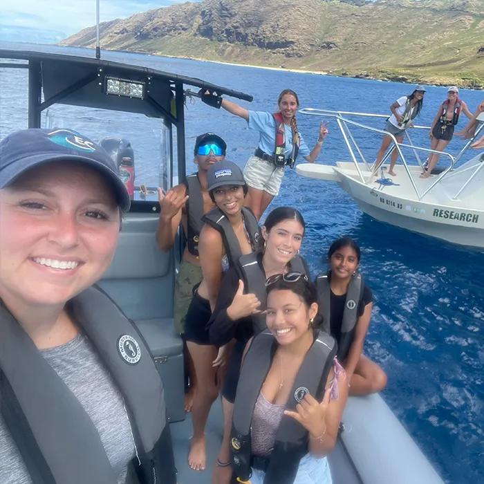 Group of smiling people in life vests on boats in blue water near rocky coastline, making hand signs.