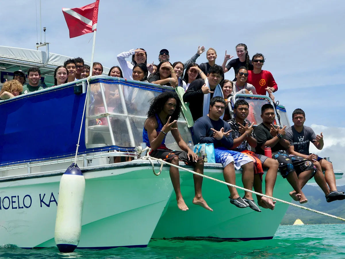 A group of people smiling and posing on a boat in clear turquoise water under a partly cloudy sky.