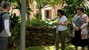 Three women stand and talk by a small stone wall in a garden with lush greenery and a building in the background.