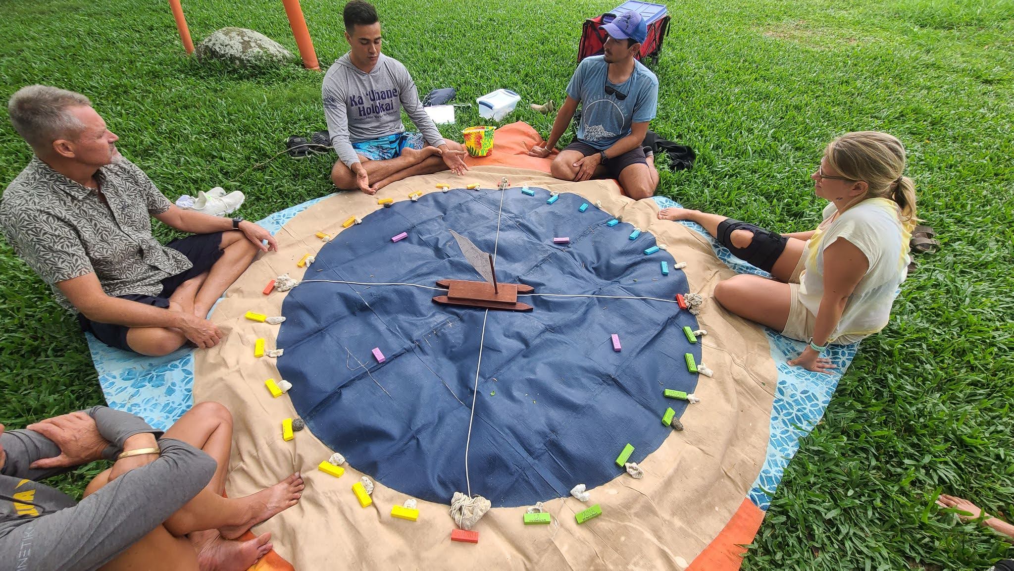 Five people sit on grass in a circle around a round game board with colored pieces and a wooden spinner.