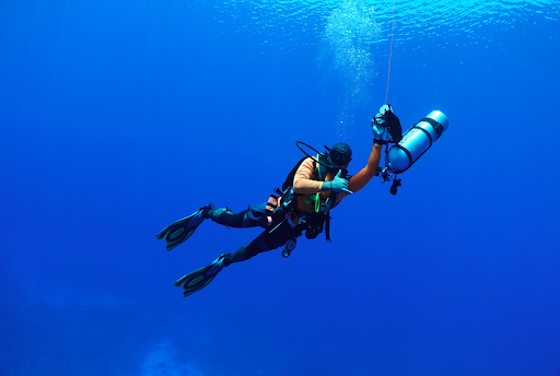 Scuba diver in deep blue water holding a line, wearing fins and carrying an air tank.