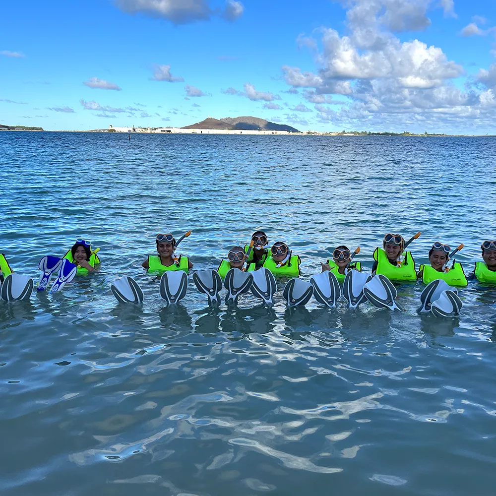 Children in bright green life jackets hold floating boards while standing in calm ocean water under a blue sky.